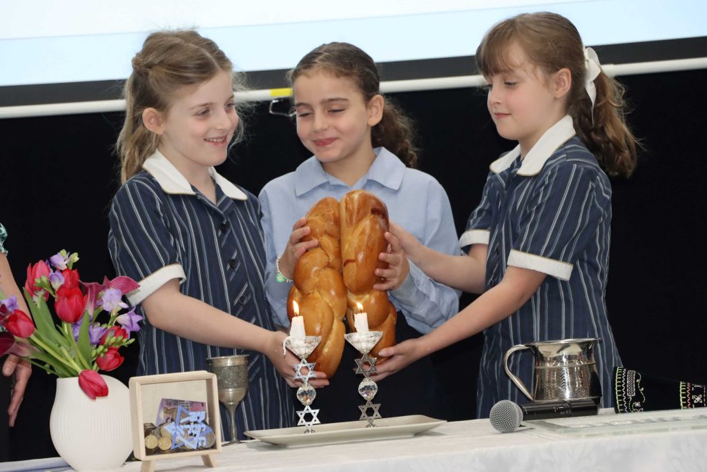 Three Year 2 girls are holding Challot, they are making the Motzi blessing in front of Shabbat chandles. They are happy.