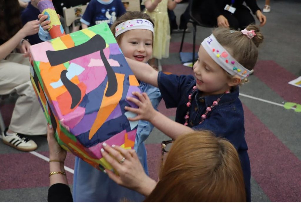 Two Junior Kinder students play with a giant dreidel-sevivon as thy celebrate Shabbat with their families.