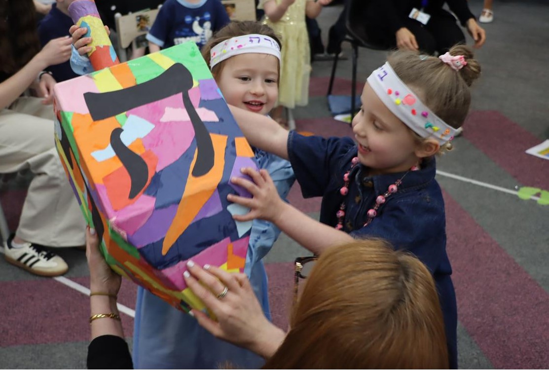 Two Junior Kinder students play with a giant dreidel-sevivon as thy celebrate Shabbat with their families.