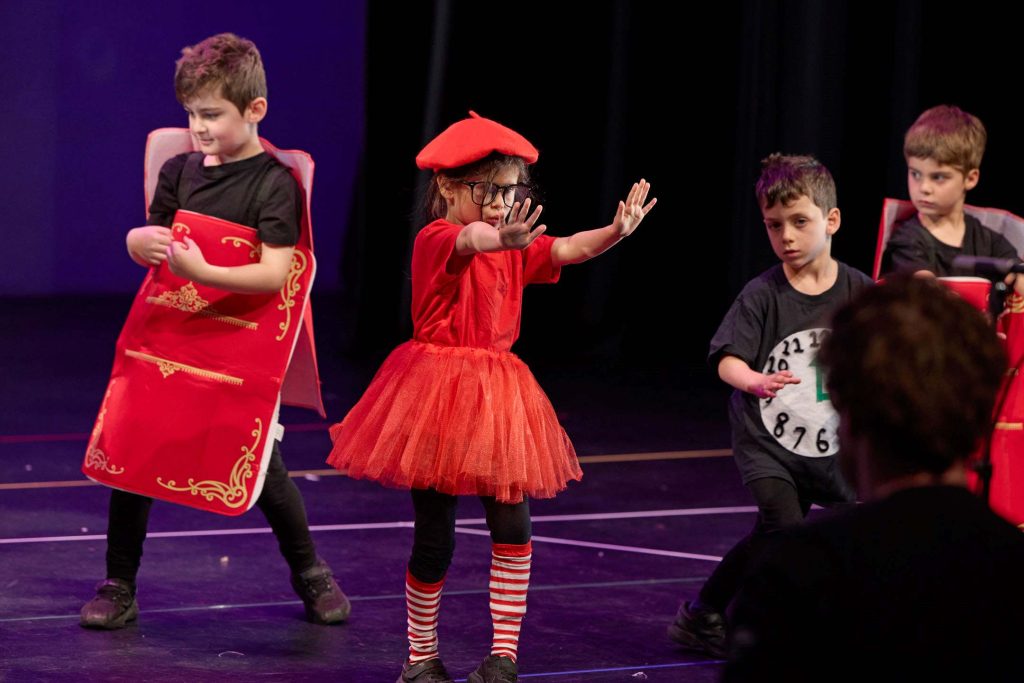 Prep to Year 2 students are in costume - one dressed as a clock, two as books, and another student is wearing a red beret and a red tutu. They are performing on stage as part of the Prep to Year 2 musical.