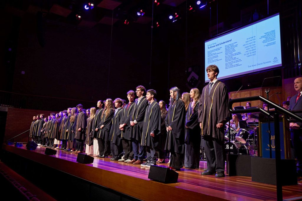The Class of 2025 stand on the stage at Robert Blackwood Hall. They are wearing graduation gowns. They are singing the School Song.