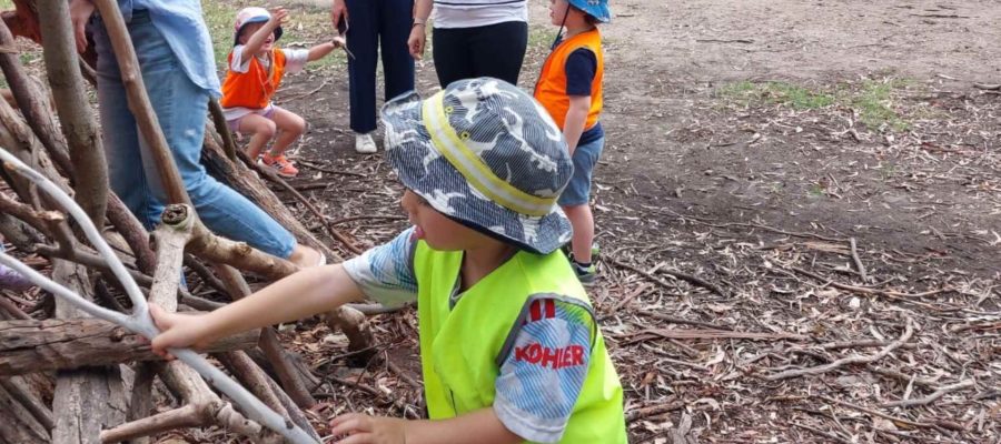 An ELC student in a high-vis vest and a hat plays with a Tee Pee made of sticks. He is at the Urban Forst Bush Kinder with his Gan Rimon KDS class.