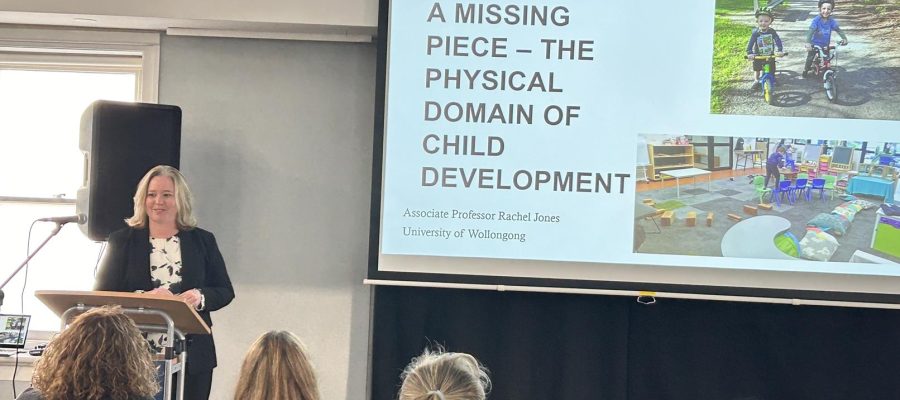 Presenter Dr Rachel Jones is speaking at a lectern. Behind her is a screen with a picture of children's playgrounds. She is presenting on the importance of movement in children's learning.