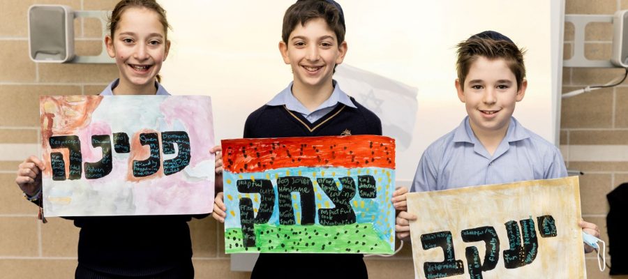 Three students are smiling looking at the camera. They hold paintings of their Hebrew names