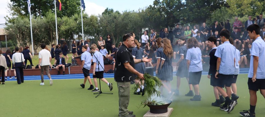 Uncle William Pepper is conducting a Smoking Ceremony to mark the start of the 2024 school year. There is smoke coming from a fire of gum leaves. Students walk past and through the smoke.