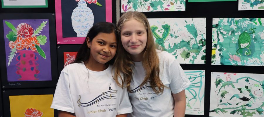 Two Year 5 students are looking at the camera and smiling. They are wearing KDS choir t-shirts. They are standing in front of colourful art painted by King David students.