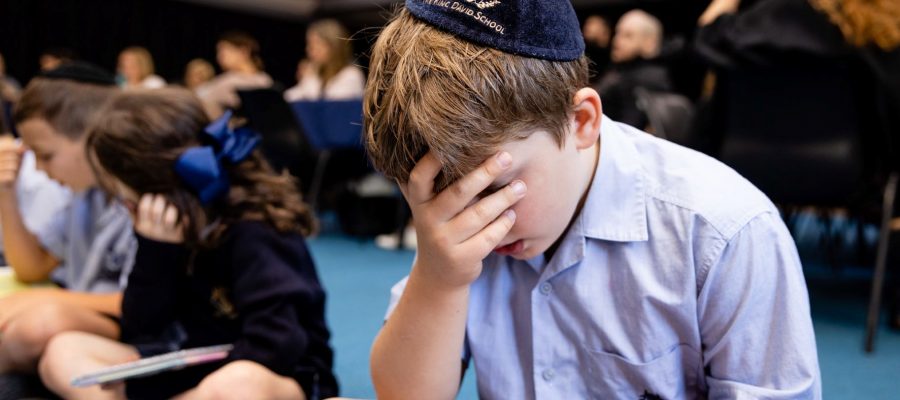 A student wears a King David School Kippah. He is covering his eyes with his hand and holding a new Siddur made for him