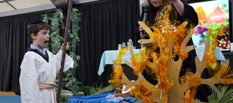 A Junior School student is dressed as Moshe when he was a shepherd. He is standing in front of a burning bush made of orange and yellow cellophane. He is about to hear God's voice.