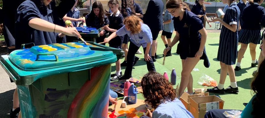 Photo shows a big rubbish bin being painted with rainbow swirls by students in uniform who are promoting sustainability