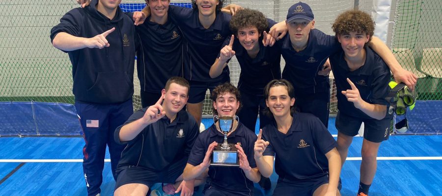 The boys soccer team is huddled together, they are looking at the camera and smiling. The boy crouching down in the middle is holding a trophy.