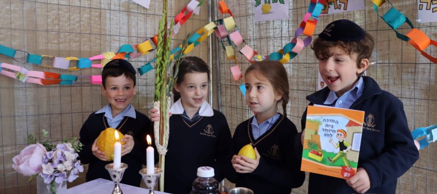 Four children are in the sukkah. Two are holding Etrogs. One holds a book in Hebrew about Sukkot,. There are Shabbat candles on the tables, along with grape juice for kiddush.