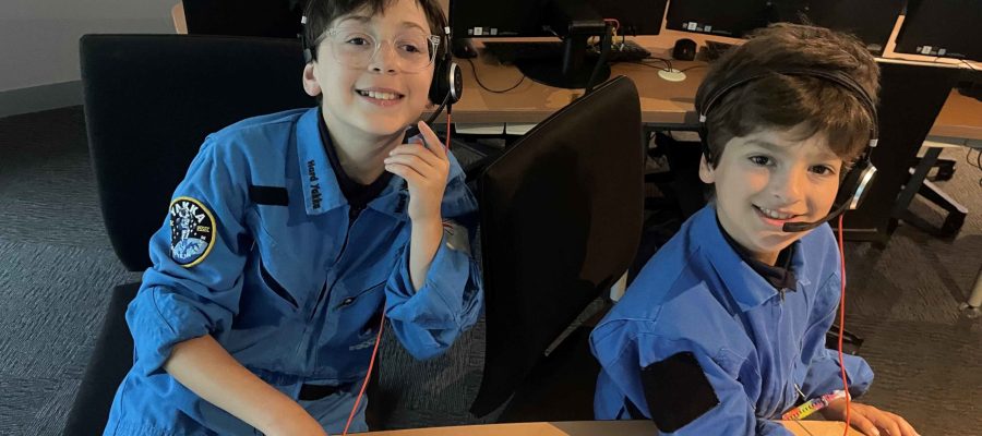 Two Year 5 students are wearing blue Space overalls, as though they work at an International Space Centre. They are wearing headsets and sitting in front of computers at the Victorian Space Science Education Centre (VSSEC)